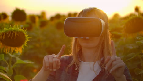 The-female-student-with-long-hair-in-plaid-shirt-and-jeans-is-working-in-VR-glasses.-She-is-engaged-in-the-working-process.-It-is-enjoyable-sunny-day-in-the-sunflower-field.